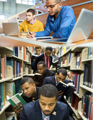 Two people sitting at a desk. Multiple people walking in between two bookshelves.